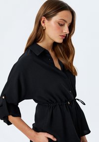 Zwarte button-up shirt met een ontspannen pasvorm, elastische taille en gouden accenten op de mouwen. De stof lijkt glad met een lichte glans.