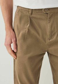 Brown cotton trousers with a textured fabric, featuring front pleats and a button closure. Side hand placed on the thigh area.