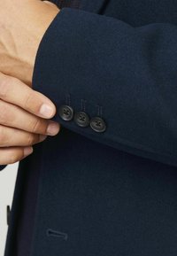 Hand som justerar ärmen på en mörkblå kappa med tre svarta knappar i en närbild av handledsområdet.