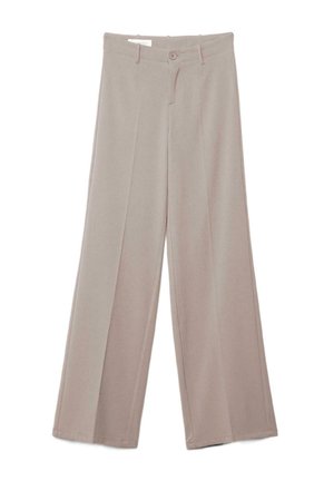 Junge Frau mit langen blonden Haaren, die ein weißes ärmelloses Oberteil, beige hoch taillierte Hose und dunkle spitze Schuhe trägt und mit verschränkten Armen steht.