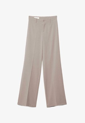 Młoda kobieta o długich blond włosach, ubrana w biały, bezrękawnik, beżowe spodnie z wysokim stanem oraz ciemne, spiczaste buty, stojąca z założonymi ramionami.