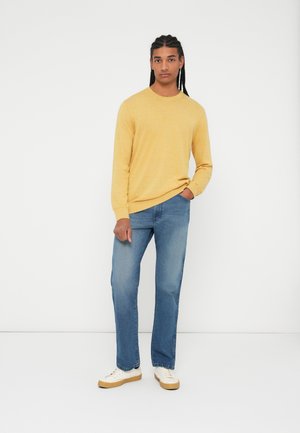 Geel langemouwtrui, blauwe rechte spijkerbroek en witte schoenen met een beige zool. Eenvoudige textuur, relaxte pasvorm, staande houding.