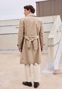 Beige trenchcoat met een tailleriem, knoopmanchetten en een rugventilatie, gecombineerd met crèmekleurige broeken en zwarte schoenen in een daktuin.