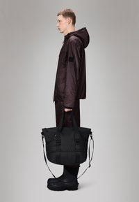 Saco de tote isolado preto com alças ajustáveis, textura suave e forma retangular, segurado por uma pessoa vestindo uma jaqueta impermeável escura.