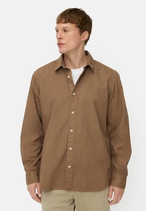 Man draagt een bruin knoopjes overhemd met lange mouwen over een wit ondershirt, kijkt naar links tegen een effen lichte achtergrond.