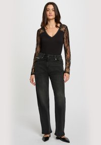 Kvinde står i en sort blonder langærmet top med gennemsigtige ærmer, højtaljede sorte jeans og spidse sorte flade sko på en hvid baggrund.