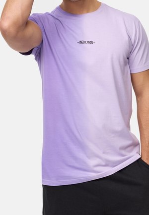Mann trägt ein hellviolettes kurzärmeliges T-Shirt mit dem Schriftzug "INDICODE" auf der Brust, schwarze Hose, und eine Hand ist nahe dem Kopf erhoben.