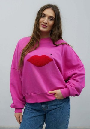 Kobieta o długich brązowych włosach, ubrana w jasnoróżową, oversize'ową bluzę z dużym wzorem czerwonych ust, w połączeniu z niebieskimi dżinsami, stojąca w zrelaksowanej pozie.