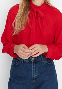 Red blouse with a tied neck, long sleeves, and gathered cuffs, paired with high-waisted blue denim jeans. Smooth fabric with a light sheen.