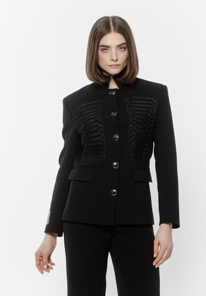 Kobieta o prostych włosach do ramion, ubrana w czarną, zapinaną na guziki kurtkę z misternym plecionym wzorem oraz dopasowane czarne spodnie na białym tle.