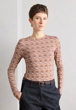 Femme aux cheveux courts portant des boucles d'oreilles en anneau doré, un haut à manches longues à motifs, un pantalon foncé et une ceinture marron, debout les mains dans les poches.