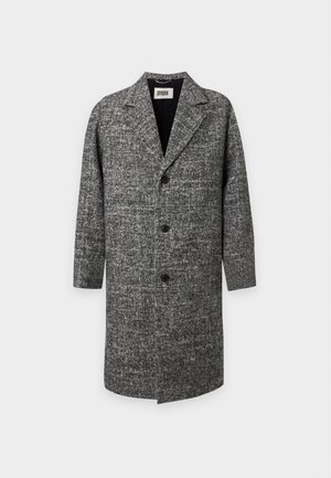 Homme portant un manteau gris texturé par-dessus une chemise sombre boutonnée, un pantalon noir, des bottes noires et un collier avec une plaque militaire sur un fond uni.