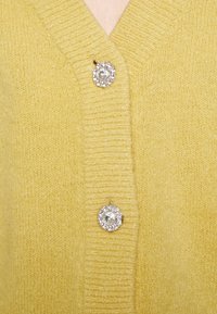Žlutý pletený cardigan se dvěma třpytivými knoflíky ve tvaru květiny upevněnými podél otevření na prsou.