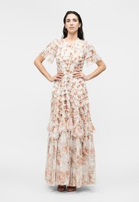 Γυναίκα που στέκεται με τα χέρια στη μέση, φοράει ένα μακρύ, βολάν, floral φόρεμα με κοντά μανίκια και φούστα με στρώσεις σε λευκό φόντο.