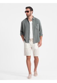 Chemise légère, boutonnée, de couleur vert sauge avec les manches retroussées sur un débardeur blanc, associée à un short crème et des chaussures slip-on beiges.