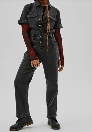 Personne portant une combinaison en denim noire avec les manches retroussées, un haut en maille rouge à manches longues en dessous, des bottes noires et une longue coiffure tressée.