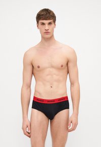 Culottes noires avec une ceinture élastique rouge portant la marque "HUGO". Le tissu semble lisse avec un design ajusté et sans motifs visibles.