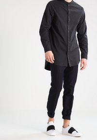Camisa de botones negra con cuello tipo banda, mangas largas y dobladillo curvado, combinada con pantalones ajustados de cuadros en tonos azul marino y zapatillas blancas sin cordones.