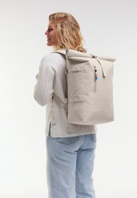 Sac à dos à rabat en beige clair. Fabriqué en matériau durable, il dispose de bretelles ajustables, d'une poche zippée sur le côté et d'accents matériels colorés.