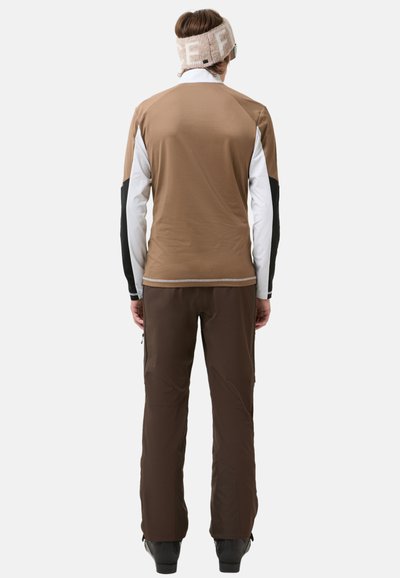 Herenoutfit voor buitenactiviteiten met een bruine, lange mouw top met witte en zwarte accenten, en bruine, slim fit broeken met ritsen aan de zijkanten.