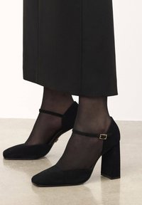 Black suede heels with a block heel, pointed toe, and strap accent. Worn with sheer black stockings, showcasing a smooth finish.