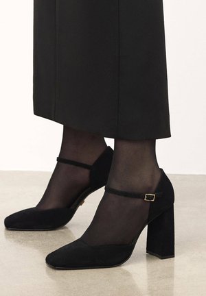 Black suede heels with a block heel, pointed toe, and strap accent. Worn with sheer black stockings, showcasing a smooth finish.