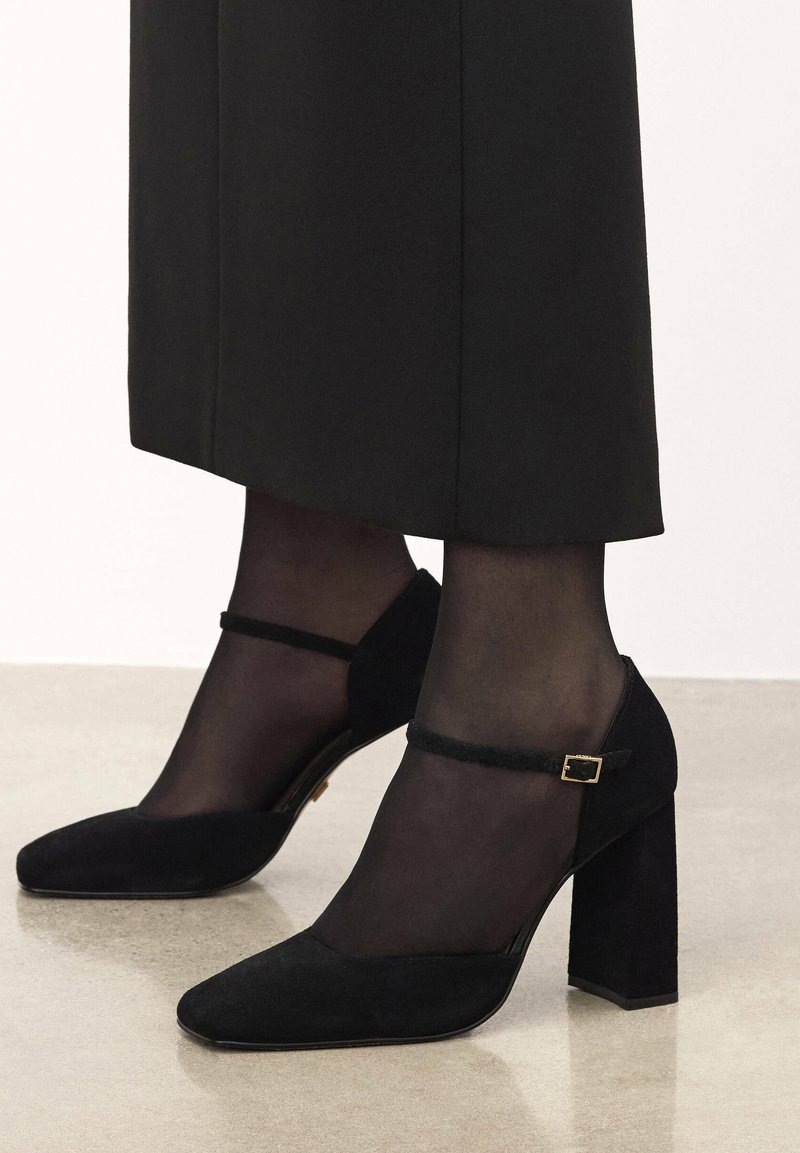 Black suede heels with a block heel, pointed toe, and strap accent. Worn with sheer black stockings, showcasing a smooth finish.