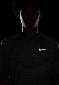 Camisa deportiva Nike de color gris oscuro con manga corta, presenta un logotipo blanco en el pecho, iluminado sobre un fondo negro.