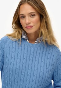 Jasnoniebieski sweter w splot warkoczowy z prążkowaną fakturą. Posiada okrągły dekolt i jest zestawiony z białą koszulą w niebieskie paski.