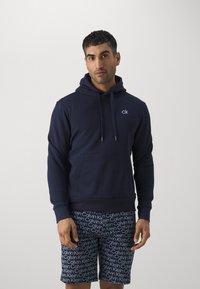 Dunkelblauer Hoodie mit einer Kordelzugkapuze, Taschen auf der Vorderseite und einem kleinen weißen Logo auf der Brust. Getragen mit gemusterten Shorts in Weiß und Blau.