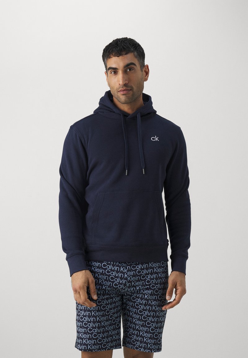 Dunkelblauer Hoodie mit einer Kordelzugkapuze, Taschen auf der Vorderseite und einem kleinen weißen Logo auf der Brust. Getragen mit gemusterten Shorts in Weiß und Blau.
