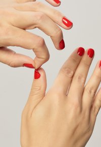 Kaksi kättä, joissa on sileä iho ja punainen kynsilakka. Toinen käsi puristaa osaa ihosta peukalon ja etusormen välissä toisessa kädessä.