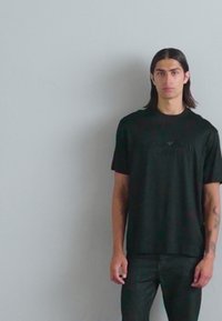 T-shirt nera in un tessuto morbido, con un logo discreto sul petto, abbinata a jeans neri, su uno sfondo grigio semplice.