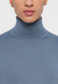 Nahaufnahme einer Person, die einen blauen gerippten Rollkragenpullover trägt, wobei der untere Gesichtsauschnitt und der Nackenbereich zu sehen sind.