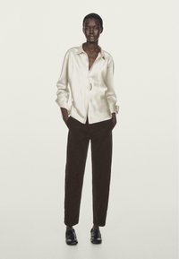 Camicia in satin avorio con vestibilità rilassata, maniche lunghe e chiusura con bottoni, abbinata a pantaloni in corduroy scuri e mocassini neri.