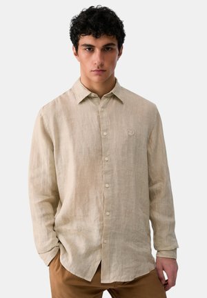 Giovane uomo con capelli ricci scuri, indossa una camicia beige di lino con bottoni e pantaloni marroni, in piedi davanti a uno sfondo chiaro uniforme.