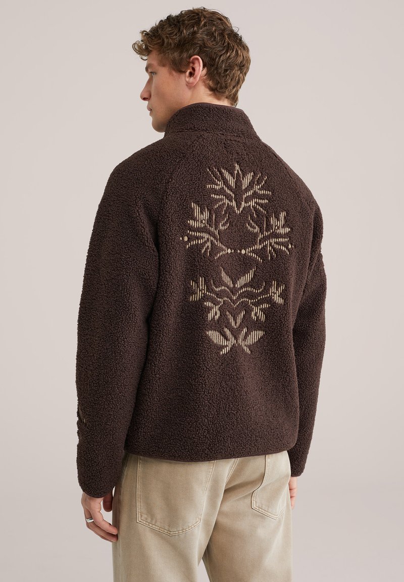 Muškarac koji nosi tamnoljubičasti runasti džemper s bež geometrijskom cvjetnom izvezenom na leđima, uz svijetlobež hlače, okrenut leđima.