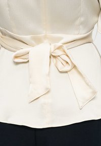 Ζώνη από ύφασμα με κρεμ υφή δεμένη σε φιόγκο γύρω από το αντίστοιχο τοπ, εμφανίζεται στο ύψος της μέσης με μαύρο παντελόνι από κάτω.