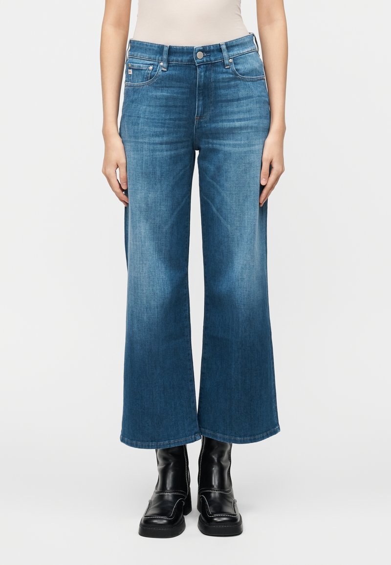 Jeans a gamba larga in denim blu con effetto sbiadito, caratterizzati da un classico design a cinque tasche e una vita media, abbinati a stivaletti neri.