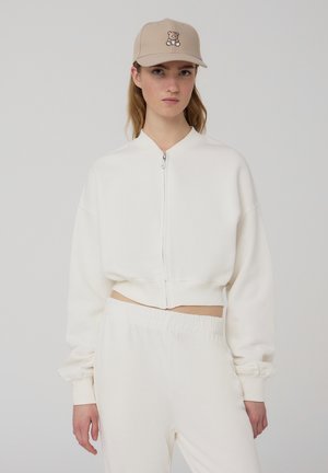 Felpa accorciata color crema con zip frontale, maniche lunghe e polsini a costine, abbinata a pantaloni della tuta coordinati. Cappello beige con design di un orso.