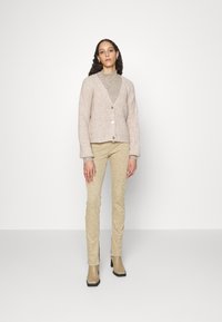 Cardigan rosa claro com lã texturizada, apresentando um decote em V e botões. Combinado com calças texturizadas bege e botas de tornozelo castanhas.