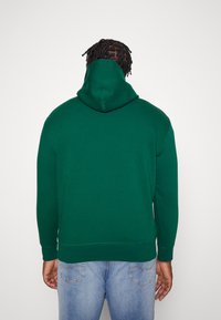 Grøn hoodie med kængurulomme, blødt stof, ribbede manchetter og kant, med hætte og et enkelt, rent design. Båret med blå jeans.
