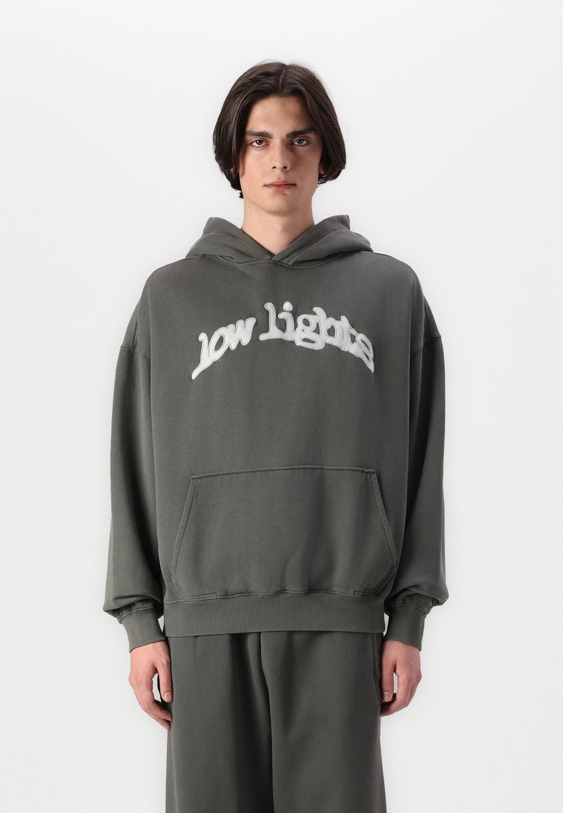 Hoodie cinza oversized com um bolso frontal, apresentando "low lights" em costura branca na parte frontal. Material de algodão suave e punhos com acabamento canelado.