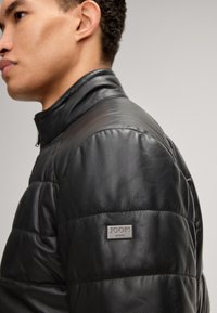 Schwarze Lederjacke mit gestepptem Textur, hohem Kragen und Metall-Logo-Patch am linken Arm. Verfügt über ein schlankes, tailliertes Design.