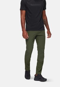 Groene outdoor broek van lichtgewicht stof, met een aansluitend ontwerp en ritszakken, gedragen met een zwart shirt met korte mouwen en donkere schoenen.