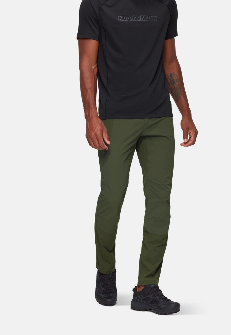 Groene outdoor broek van lichtgewicht stof, met een aansluitend ontwerp en ritszakken, gedragen met een zwart shirt met korte mouwen en donkere schoenen.