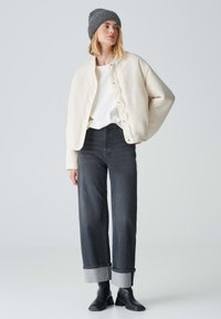 Veste bomber couleur crème avec des boutons-pression, portée sur une chemise blanche. Jeans noirs avec les poignets retroussés et bottines noires. Bonnet gris.