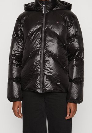 Doudoune noire avec capuche, finition brillante, motif matelassé diagonal et fermeture éclair frontale. Portée par-dessus un pantalon noir.