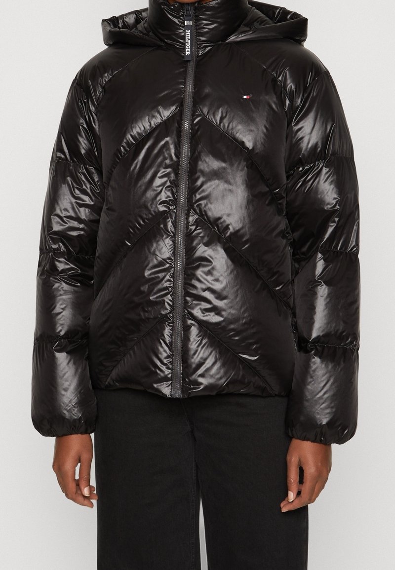 Doudoune noire avec capuche, finition brillante, motif matelassé diagonal et fermeture éclair frontale. Portée par-dessus un pantalon noir.