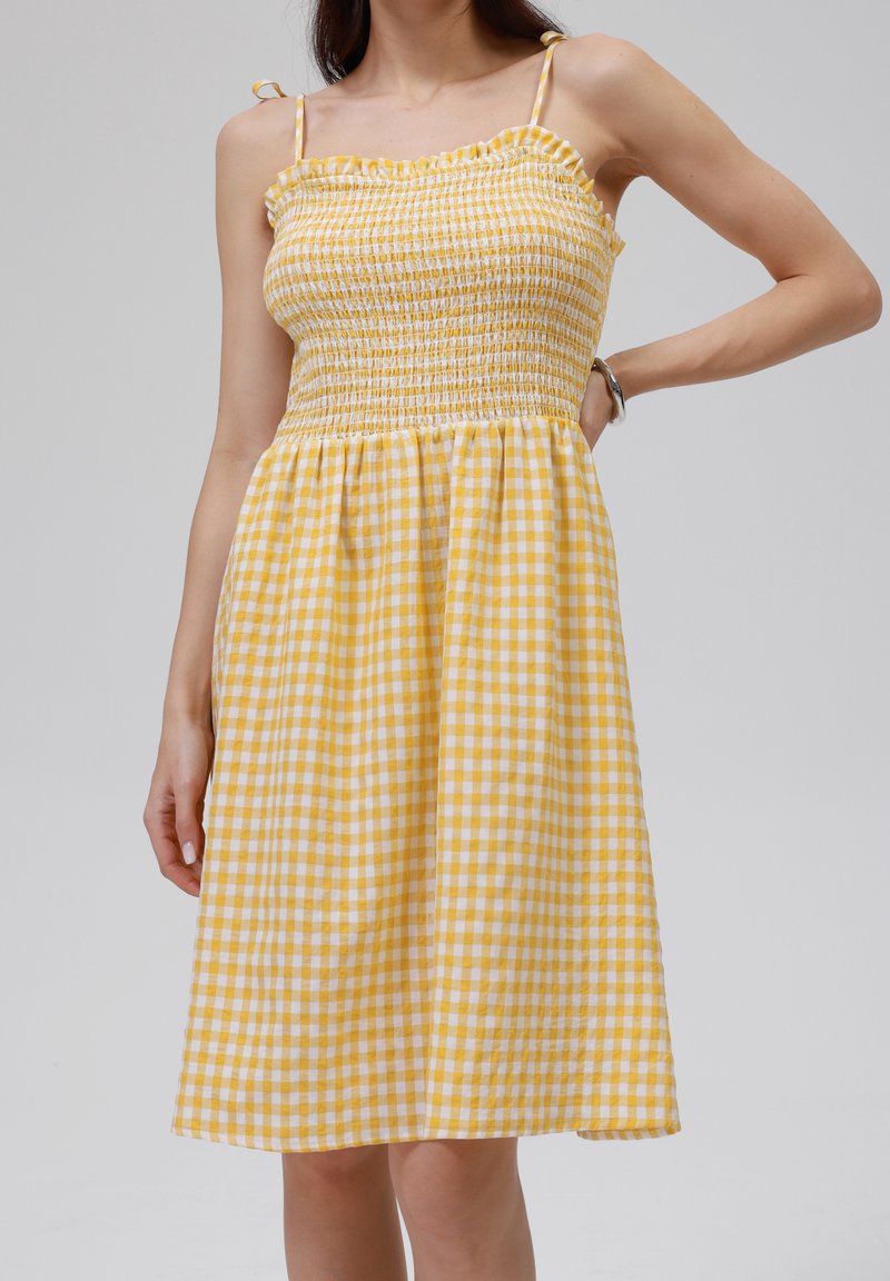 Žluté kostkované šaty s vyšívaným živůtkem, volánkovým výstřihem a nastavitelnými ramínky. Materiál je lehký s jemnou texturou.